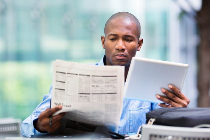 Persona seduta che legge un giornale finanziario mentre tiene un tablet nella mano sinistra.